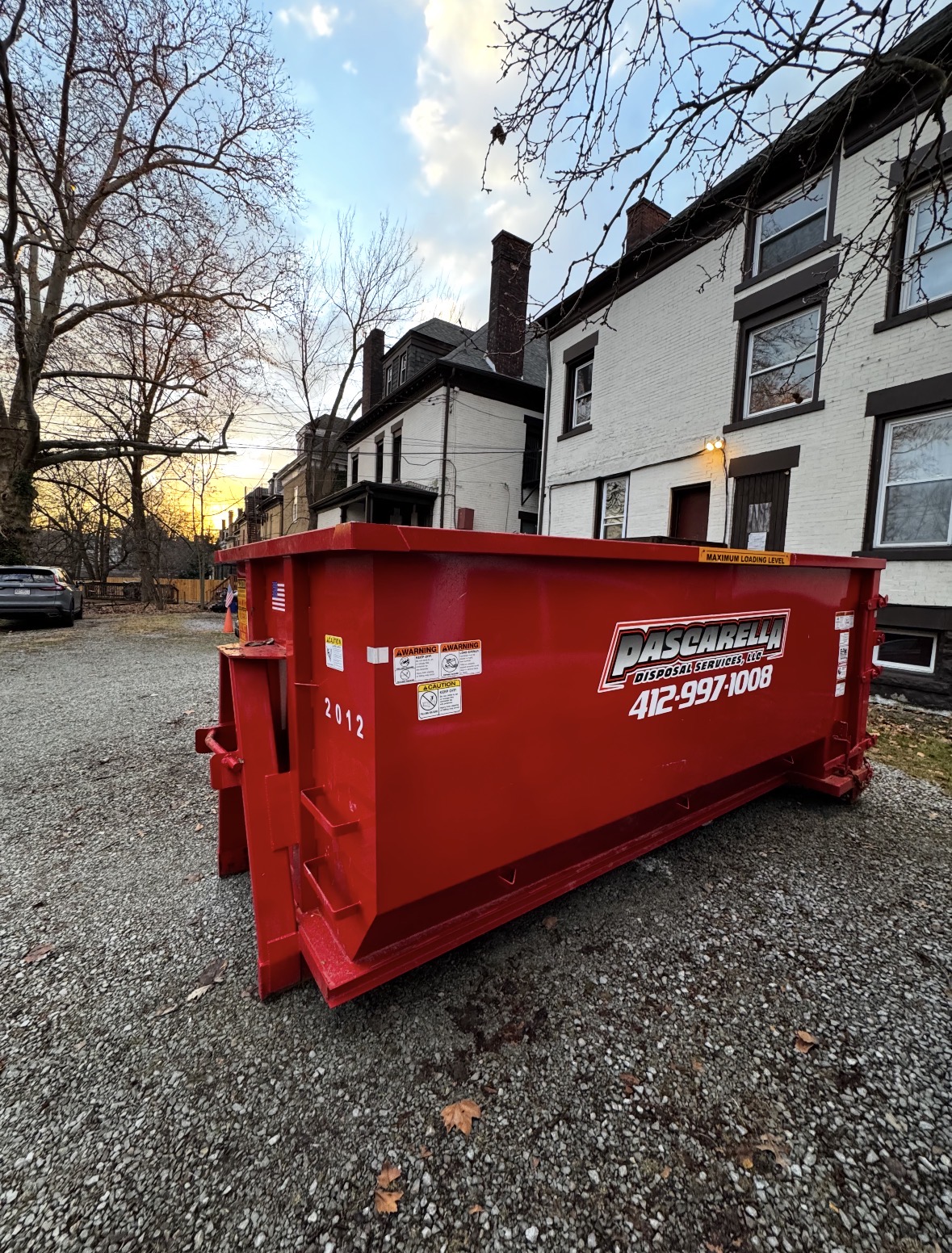 Freshly painted Pascarella dumpster in a gravel courtyard beside Pittsburgh rowhomes at sunset