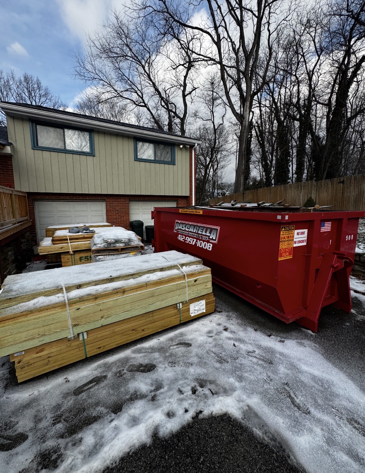 Pascarella dumpster on a snowy driveway alongside stacks of fresh lumber