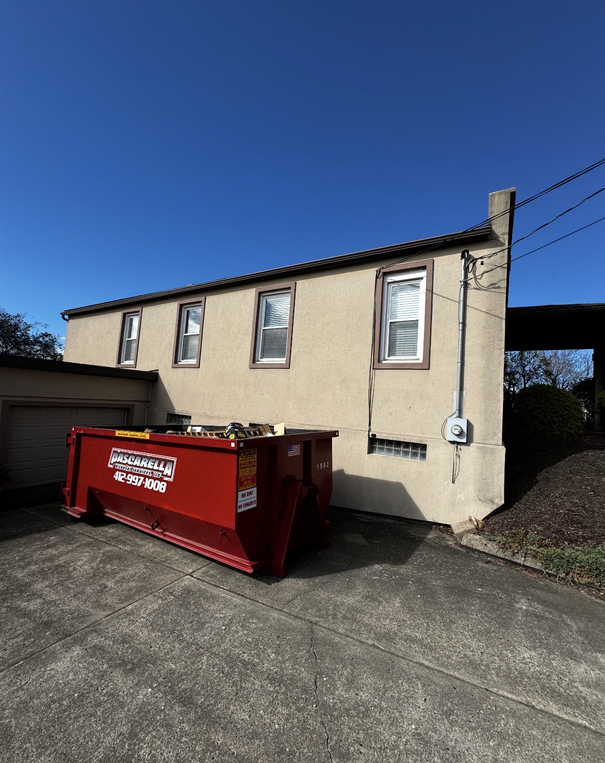 Pascarella dumpster staged on a sunny driveway beside a beige home
